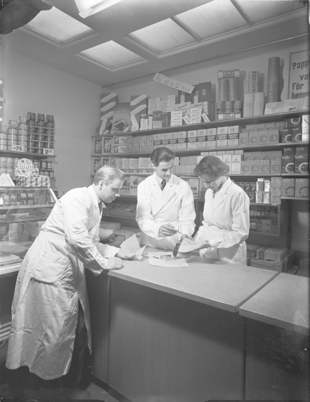 Drei Männer in weißen Kitteln an einer Apotheken-Theke mit Papieren und Gegenständen in einem schwarz-weißen Setting mit Regalen im Hintergrund.