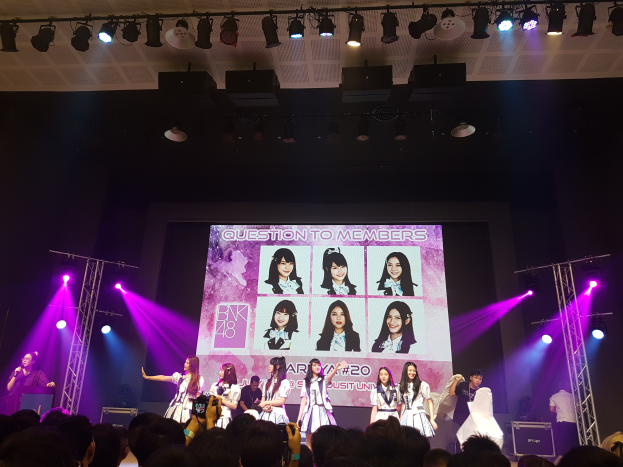 Eine Gruppe von Menschen, die auf einer hell erleuchteten Bühne performen, mit einer Menge darunter, einem Menschen mit Mikrofon und einem anderen, der ein Instrument spielt, und einem großen Bildschirm im Hintergrund, der Bilder von BK48 zeigt.