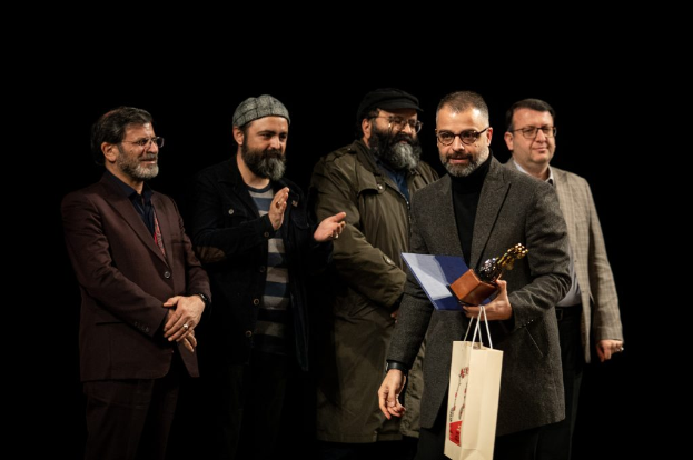 Eine Gruppe von Männern steht auf einer Bühne, einer hölt eine Tasche und eine Trophäe, mit einem dunklen Hintergrund, der die Eröffnung der Berliner Filmfestspiele suggeriert.