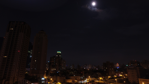 Eine nächtliche Stadtansicht mit hohen Gebäuden, Häusern und zahlreichen Straßenlaternen unter einem Himmel mit sichtbarem Mond.