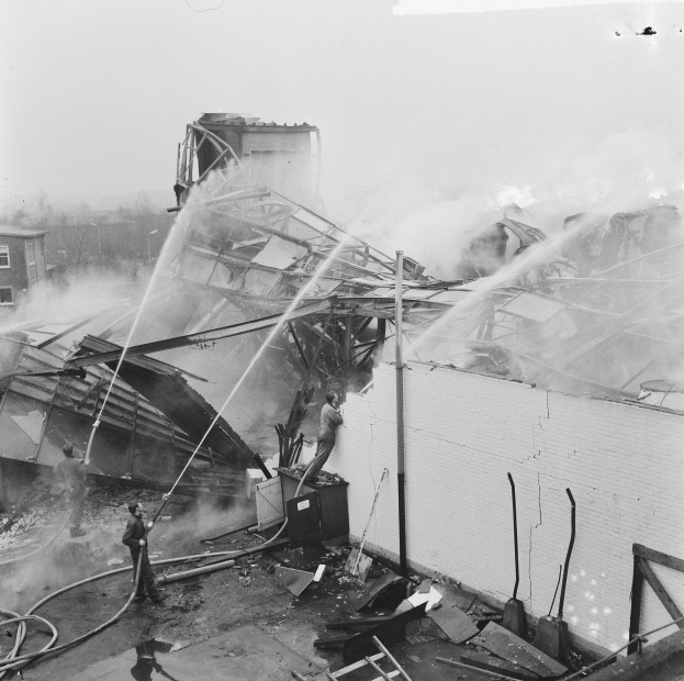 Schwarzes und weißes Foto von Feuerwehrmännern auf einer Leiter, die einen Dachbrand bekämpfen, während Zuschauer unten Ausrüstung halten.