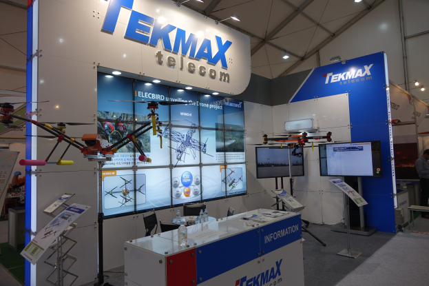 Ein Messestand für Tekmax Telecom mit einer großen Wandanzeige, einem Tisch mit Flaschen und Gegenständen, zwei Fernsehern, Texttafeln, Deckenleuchten und einigen stehenden und sitzenden Personen.