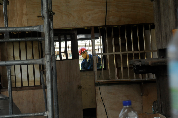 Ein Mann mit rotem Helm steht vor einem Fenster, daneben sind eine Tür, eine Leiter und eine Flasche zu sehen.