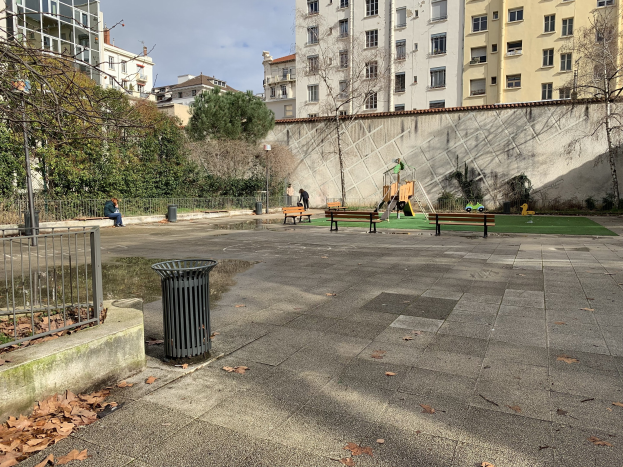 Ein Park mit Bänken, einem Mülleimer, trockenen Blättern, einem Metallzaun, Bäumen, Pflanzen, einer Wand, Pfosten, einem Straßenpfosten, Menschen auf den Bänken sitzend, Kindern auf der Wiese spielend, Gebäuden mit Fenstern und einem bewölkten Himmel.