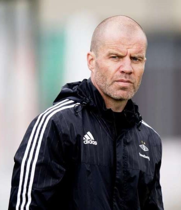 Baldiger Trainer in marineblauer Jacke mit weißen Streifen und weißem Kragen auf einem Fußballfeld mit verschränkten Händen und ernstem Gesichtsausdruck.