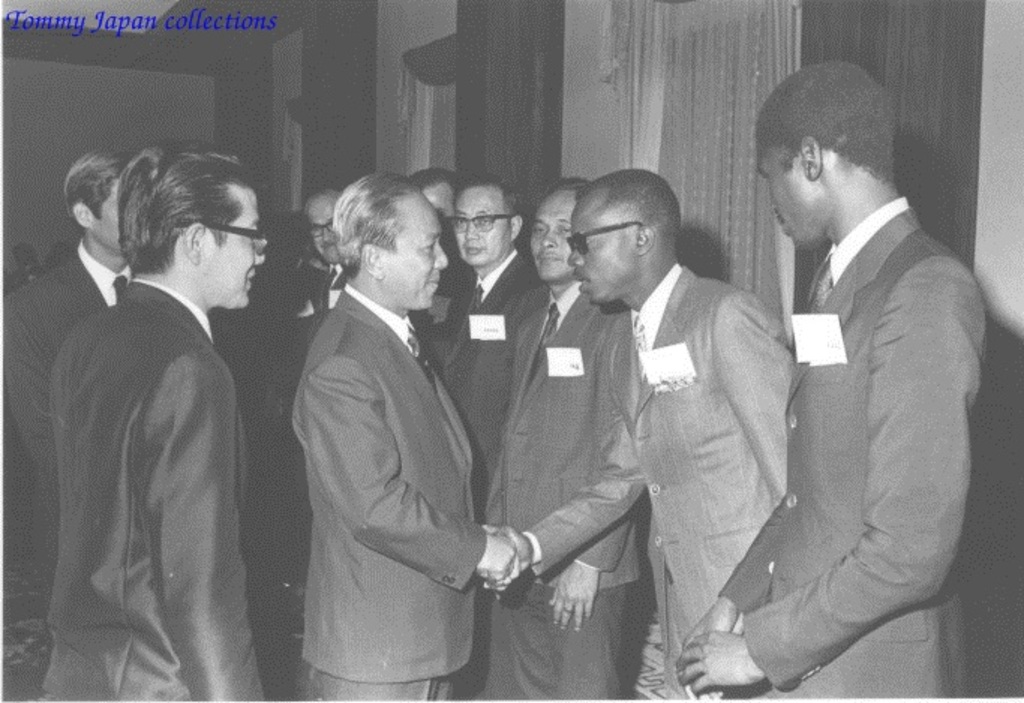 Ein Schwarz-Weiß-Bild von Männern in Anzügen, die vor Vorhängen und einer Wand stehen, mit einem Wasserzeichen im linken oberen Eck.