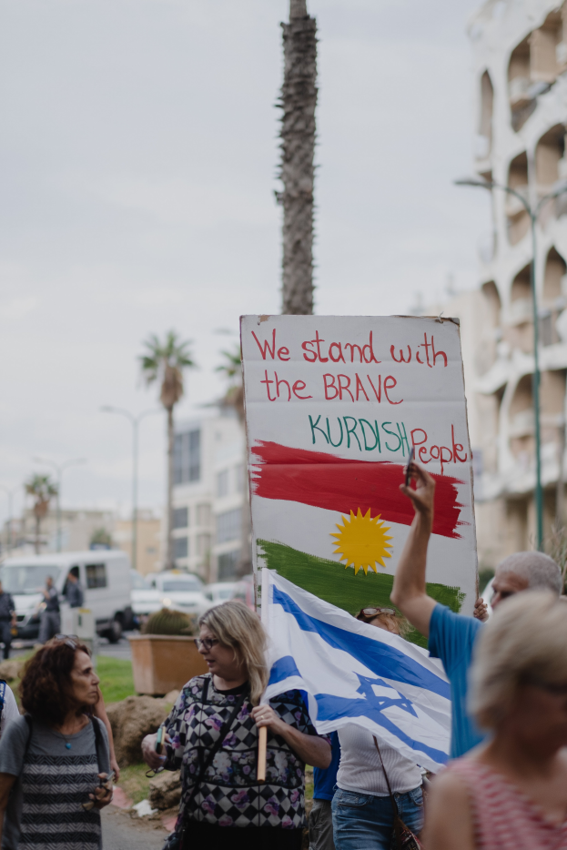 Eine Gruppe von Menschen marschiert nachts eine Straße entlang, mit einer Tafel, auf der steht: "Wir stehen zu den tapferen kurdischen Menschen" im Hintergrund ein geparkter Van, Straßenlaternen, Bäume und Gebäude.
