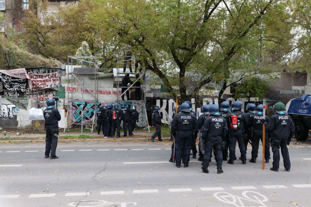 Polizisten in schwarzer Uniform und Helmen stehen vor einem Graffiti-bedeckten Gebäude, einige halten Stöcke, mit Bäumen, Gebäuden, einem klaren Himmel und einem geparkten Fahrzeug im Hintergrund.