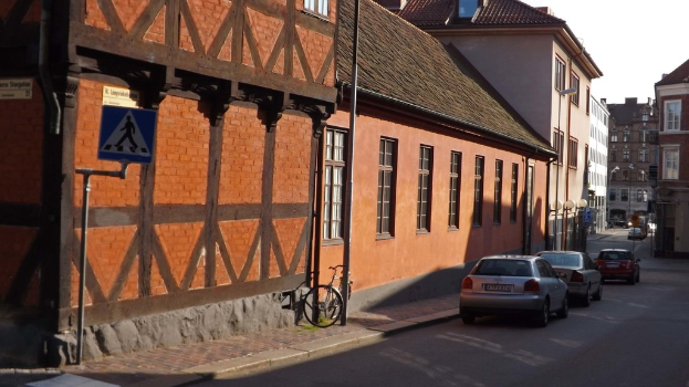 Fahrzeuge auf der Stra√üe mit Geb√§uden, Laternen, Verkehrsschildern, Fenstern und D√§chern im Hintergrund und ein Fahrrad auf dem Gehweg in der N√§he eines Pfahls.