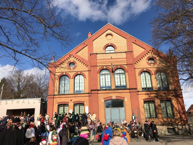 Eine große Gruppe von Menschen steht vor einem mehrstöckigen Gebäude mit Fenstern und Türen im Freien, umgeben von Bäumen unter einem bewölkten Himmel, wahrscheinlich bei einem Stadtfest.