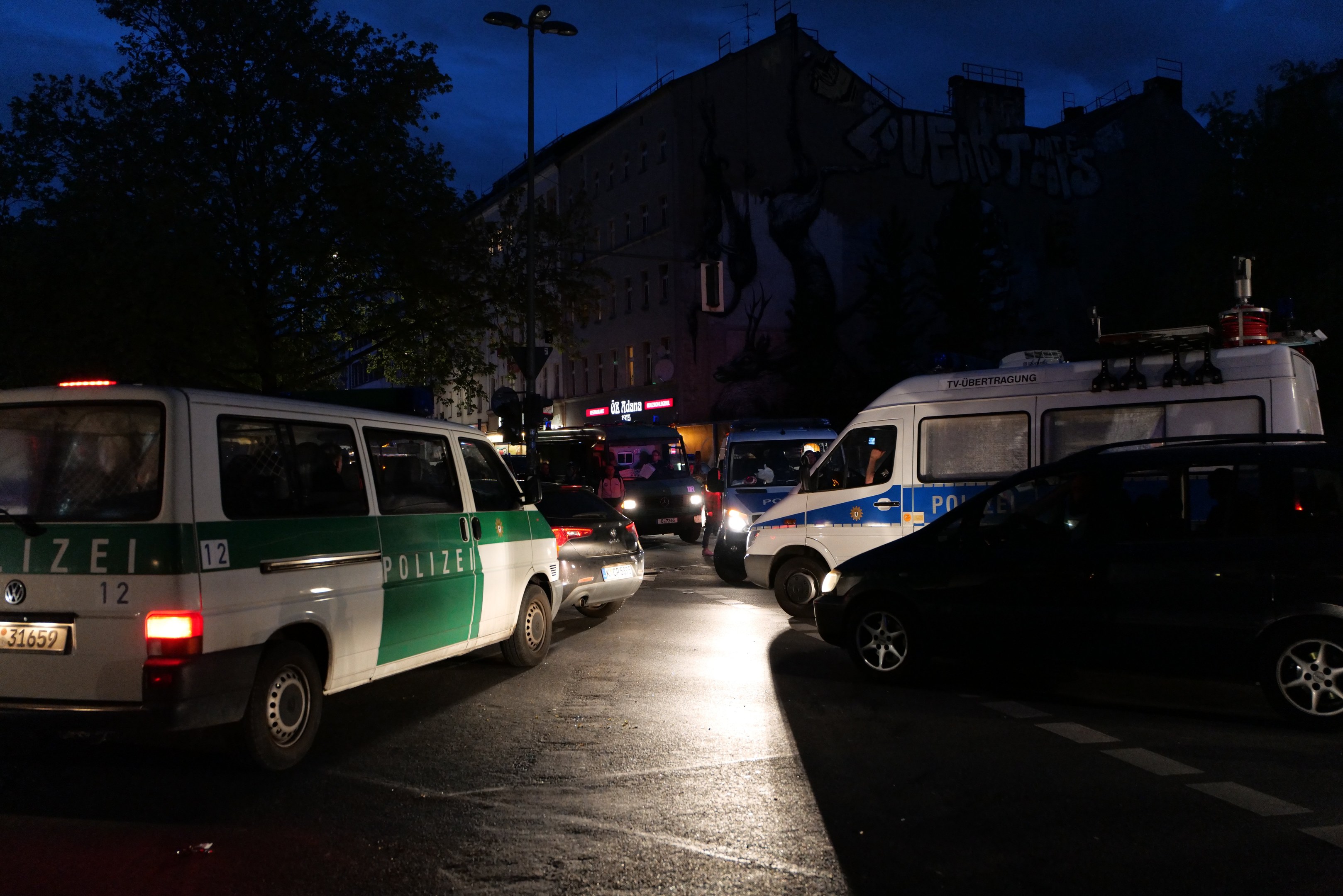 Eine Gruppe von Polizeiwagen, darunter ein Van, ein Auto und ein Rad, geparkt am Straßenrand bei Nacht mit Gebäuden, Bäumen, Laternenpfählen und einem sternenklaren Himmel im Hintergrund.