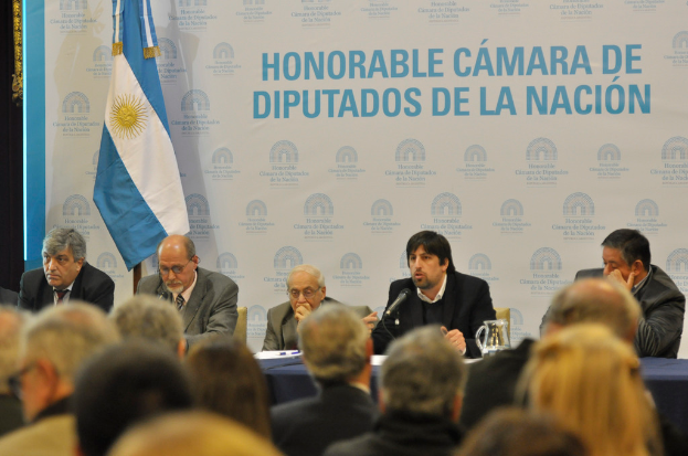 Eine Gruppe von Menschen sitzt in Reihen von Stühlen, mit einem Tisch vor der ersten Reihe, der einen Krug, Papiere und einen Stift enthält. Eine Person in der Mitte spricht in ein Mikrofon, mit einer weißen und blauen Flagge dahinter und einem weißen und blauen Hintergrund, auf dem 'Ehrenwerte Camara De' angezeigt wird.