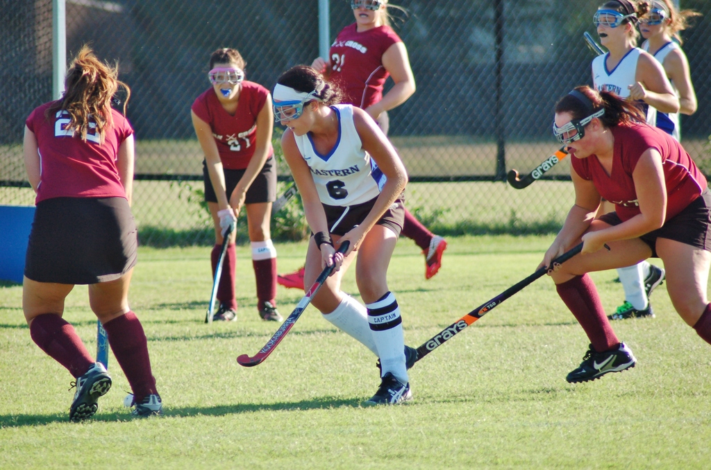 Frauen spielen Hockey auf einem Rasenfeld mit einem Maschendraht-Hintergrund.