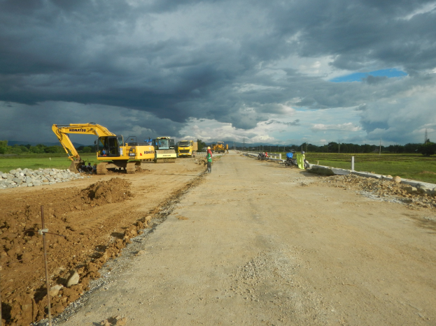 Schotterstraße mit Baumaschinen wie Kränen, Steinen, Pfählen und Menschen; Bäume, Gras und bewölkter Himmel im Hintergrund.