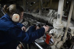 Zwei Frauen in blauen Uniformen arbeiten an einer Maschine in einer Öl- und Gasindustrieumgebung, wobei eine ein Objekt hält und Kopfhörer trägt, und im Hintergrund Rohre zu sehen sind.