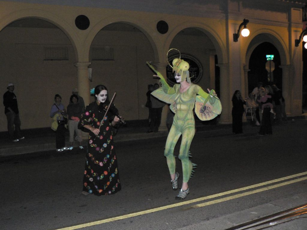 Eine Person in einem Kostüm steht neben einer Frau, die eine Geige spielt, mit Menschen und einem beleuchteten Gebäude im Hintergrund und einer Straße unten.