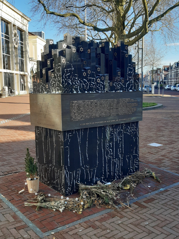 Denkmal in einem Stadtplatz für Holocaust-Opfer, mit einem Baum davor, einem Topfgewächs am Boden, verstreuten trockenen Blättern und Gebäuden, Fahrzeugen, Pfählen und Bäumen im Hintergrund.