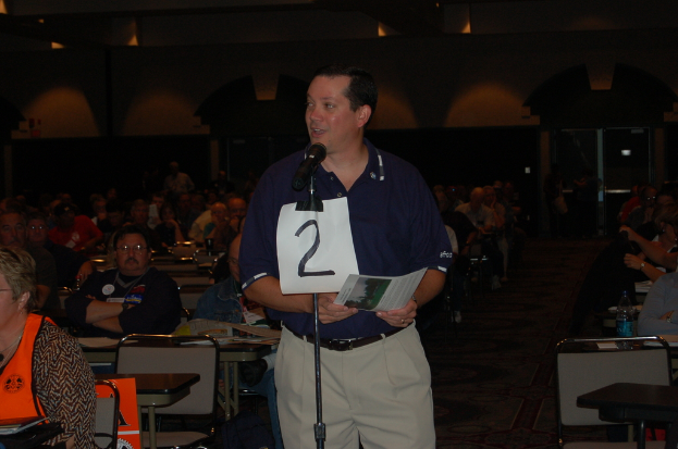 Innenansicht eines Stadions, ein Mann in einem blauen T-Shirt spricht in ein Mikrofon auf einem Ständer, hält ein Blatt Papier in der Hand, mit Gruppen von Menschen links und rechts auf Stühlen sitzend, eine Wasserflasche und ein Buch auf einem Tisch und eine Person in der Nähe einer Tür im Hintergrund.