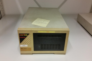 Ein cremefarbener und schwarzer elektronischer Apparat, ein Solarstrom-DDS-10, steht auf einem weißen Tisch mit einem daran befestigten Blatt Papier, vor einer weißen Wand.