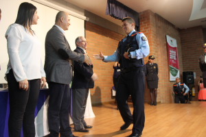 Eine Gruppe von Menschen auf einer B√ľhne, einer in Uniform mit einer M√ľtze haltend, eine Frau in einem wei√üem Hemd, mit einem Banner, Lautsprecher, Vorh√§ngen, Tisch und Wandmonitor im Hintergrund.