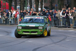 Ein grüner Wagen fährt auf einer von Geländern, Bäumen und Gebäuden gesäumten Straße an einer Menge vorbei, einige tragen Möwen und halten Kameras, mit Schildern auf Ständen im Hintergrund.