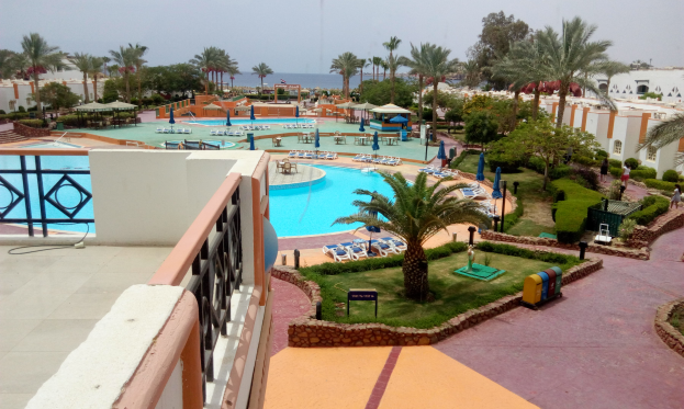 Ein Resortblick von einem Balkon aus, der einen Swimmingpool umgeben von Palmen, Gras und Pflanzen zeigt, mit einigen Menschen und einem klaren blauen Himmel im Hintergrund.