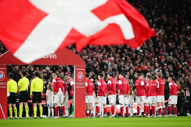 Gruppe von Menschen auf einem Fußballfeld mit einer roten und weißen Flagge im Vordergrund, einem Bogen mit der Aufschrift "Bayern München vs Bayern München Wetten & Vorschau" im Hintergrund und einer großen Menschenmenge im Stadion.