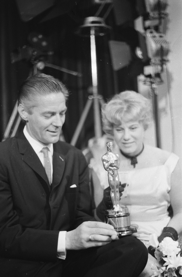 Ein Mann in einem schwarzen Blazer, weißem Hemd und Krawatte hält eine Oscar-Statue in der Hand und sitzt neben einer Frau, die einen Blumenstrauß hält, beide in Abendgarderobe, mit Lichtern im Hintergrund, in Schwarz-Weiß.