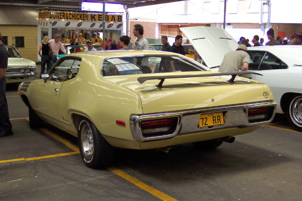 Ein Oldtimer fahrt auf einer Strasse mit Menschen am Rand, Gesch├Ąften in der Mitte und Geb├Ąuden im Hintergrund.
