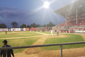Baseballspiel im Gange mit Zuschauern auf den RÀngen, BÀumen, Masten und Stadionlampen gegen einen klaren blauen Himmel.