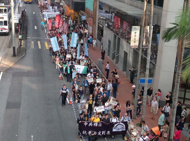 Eine große Gruppe von Menschen marschiert auf einer Demonstration durch die Straßen von Hong Kong, hält Schilder und Plakate hoch, mit Bäumen, gläsernen Gebäuden, Fahrzeugen und Straßenschildern im Hintergrund.