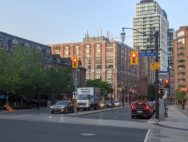 Eine belebte Stadtstraße mit verschiedenen Fahrzeugen, darunter ein Lastwagen, ein Auto und ein SUV, umgeben von Laternenmasten, Ampeln, Texttafeln, Bäumen, Gebäuden mit Fenstern unter einem Himmel.