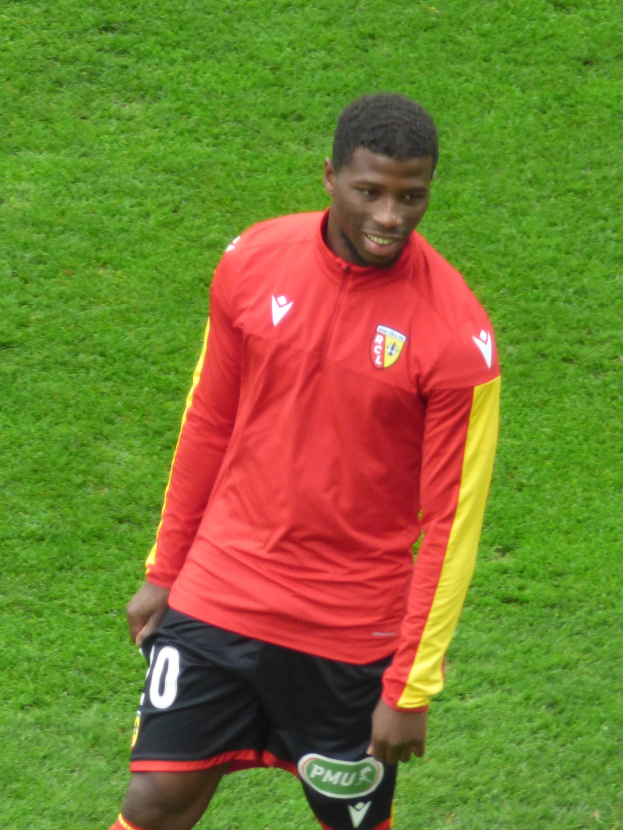 Ein Mann in rotem T-Shirt und schwarzen Shorts steht auf einem Fußballfeld, lächelt und ist bereit, als Flügelspieler zu spielen.