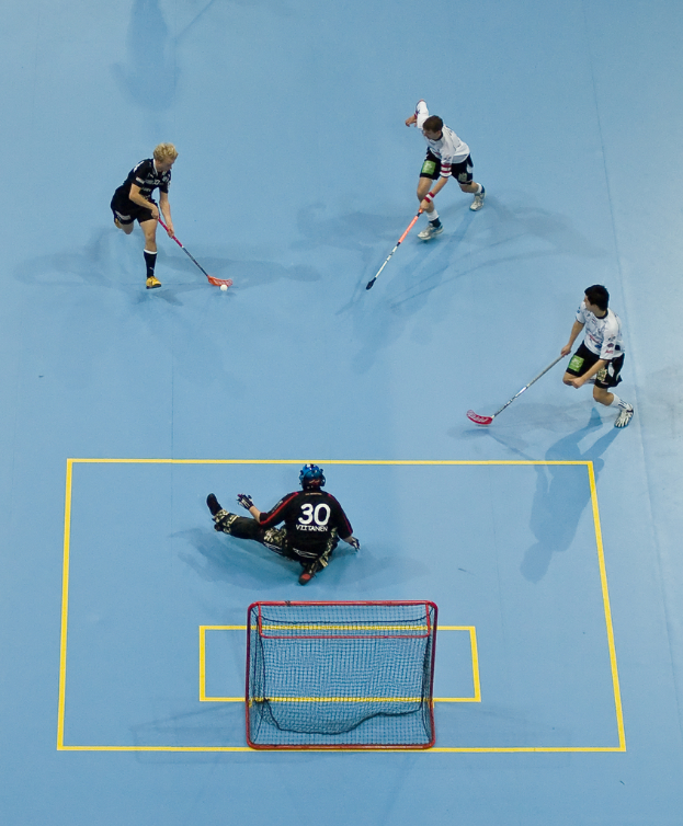 Drei Personen in Sportkleidung spielen Hockey, wobei eine Person rechts mit einem Hockeystock einen Ball schlägt und eine weitere Person hinter einem Netz auf dem Boden sitzt.