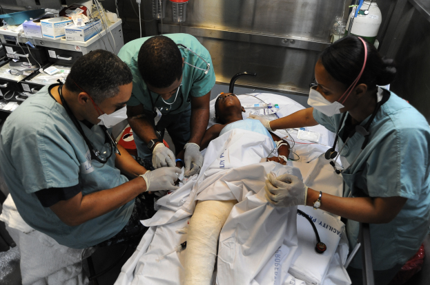 Eine Gruppe von vier Personen in Kitteln und Masken versorgt einen Patienten auf einer Trage in einem Krankenhauszimmer, mit medizinischer Ausrüstung und einem Tisch mit Gegenständen im Hintergrund.