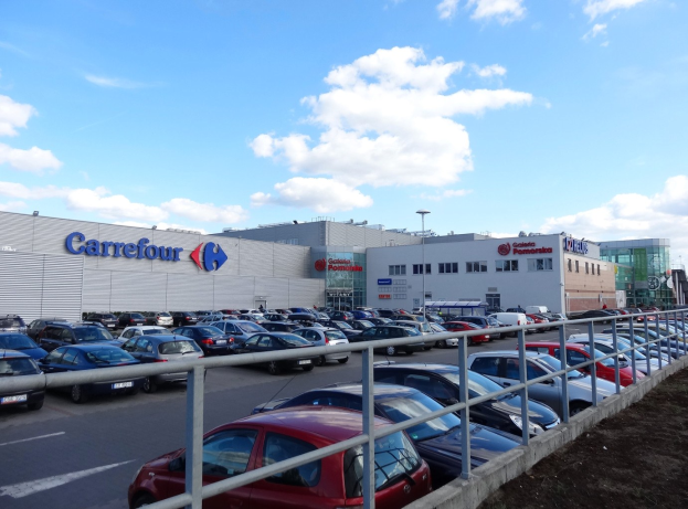 Großes Autohaus mit parkenden Fahrzeugen, umgeben von städtischer Infrastruktur wie Gebäuden, Straßenlaternen und Fahrzeugen auf der Straße unter einem bewölkten Himmel.