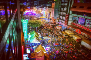 Eine bunte Faschingszene mit einer großen Menschenmenge auf der Straße, umgeben von Zelten, Geschäften, Gebäuden und Bannern, mit zahlreichen Lichtern im Hintergrund.