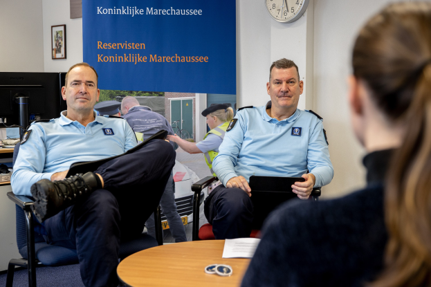 Zwei Polizeibeamte sitzen an einem Tisch mit Papieren, unterhalten sich mit einer Frau; eine Wand mit einem Banner, einer Uhr und Fotorahmen im Hintergrund und ein weiterer Tisch mit einem Monitor, Tastatur und Maus links daneben.