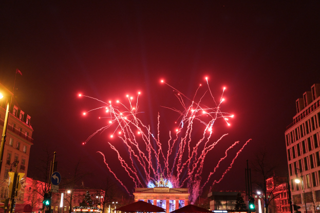 Eine Straßenszene in einer Stadt mit Gebäuden, Bäumen, Laternen, Verkehrszeichen, Zeltplanen und Menschen, mit Feuerwerk, das den Himmel erhellt, während des Neujahrsfestes in Berlin.