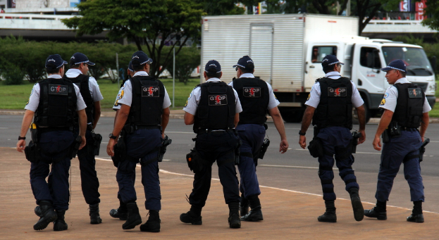 Eine Gruppe von Beamten in Uniform, einschließlich Mützen, Hemden, Schutzwesten, Uhren, Hosen und Schuhen, geht auf einer Straße. Rechts daneben steht ein Lastwagen in der Nähe eines Zauns, mit Gebäuden, Bäumen, Pflanzen, Pfählen und Bannern im Hintergrund und einer Brücke oben rechts.