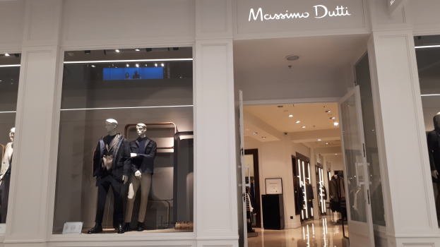 Innenansicht des Massimo Dutti-Ladens in Mailand mit Schaufensterpuppen in zeitgemäßer Kleidung, beleuchteten Deckeninstallationen, Informationsschildern und sichtbarem Bodenbelag.