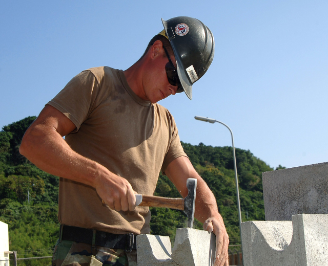 Ein Mann in einem Helm und einer Schutzbrille baut eine Wand mit einem Hammer, mit Bäumen, einem Laternenmast und einem klaren blauen Himmel im Hintergrund.