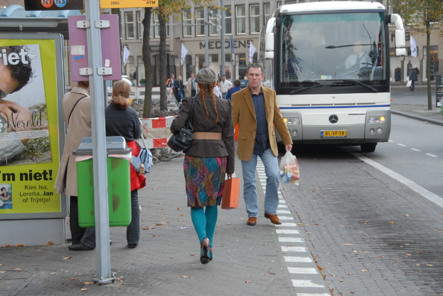Eine Gruppe von Menschen, die auf einem Fußweg neben einem silbernen Bus gehen, mit einem braunen Gebäude mit Glasfenstern im Hintergrund und einer weißen und grünen Bannerwand auf der linken Seite.