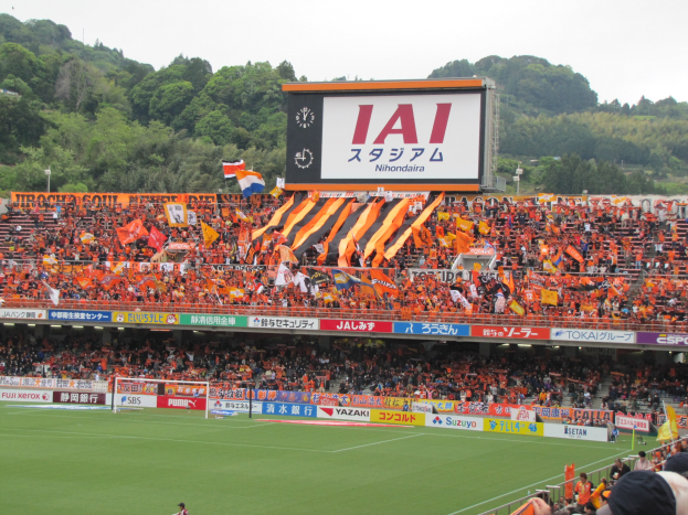 Fußballspiel in einem Stadion mit großer Zuschauermenge, saftigem Grün, einem Tor, Bannern, Fahnen, einem großen Bildschirm, Bäumen und einem klaren blauen Himmel.