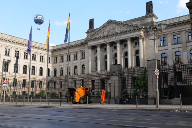 Das Reichstagsgebäude in Berlin, Deutschland, ein großes Gebäude mit zahlreichen Fenstern, umgeben von Straßenelementen, Fahrzeugen, Menschen und Bäumen, unter einem bewölkten Himmel.
