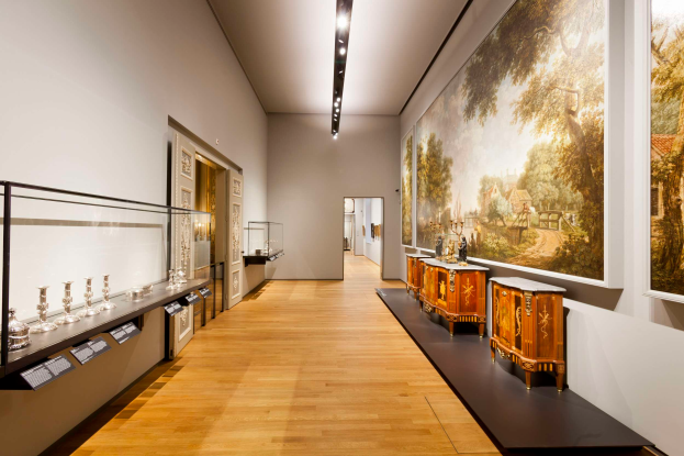 Ein Raum im Stedelijk Museum in Amsterdam, der verschiedene Kunstwerke, einschließlich Gemälde, Objekte, Texttafeln und eine Glasbox mit Gegenständen darin, zeigt, beleuchtet von Deckenlampen.