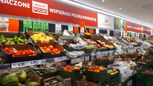 Ein Geschäft mit verschiedenen Obst- und Gemüsesorten in Kartonschachteln mit Preisschildern, Informationsschilder an den Wänden und Deckenleuchten.