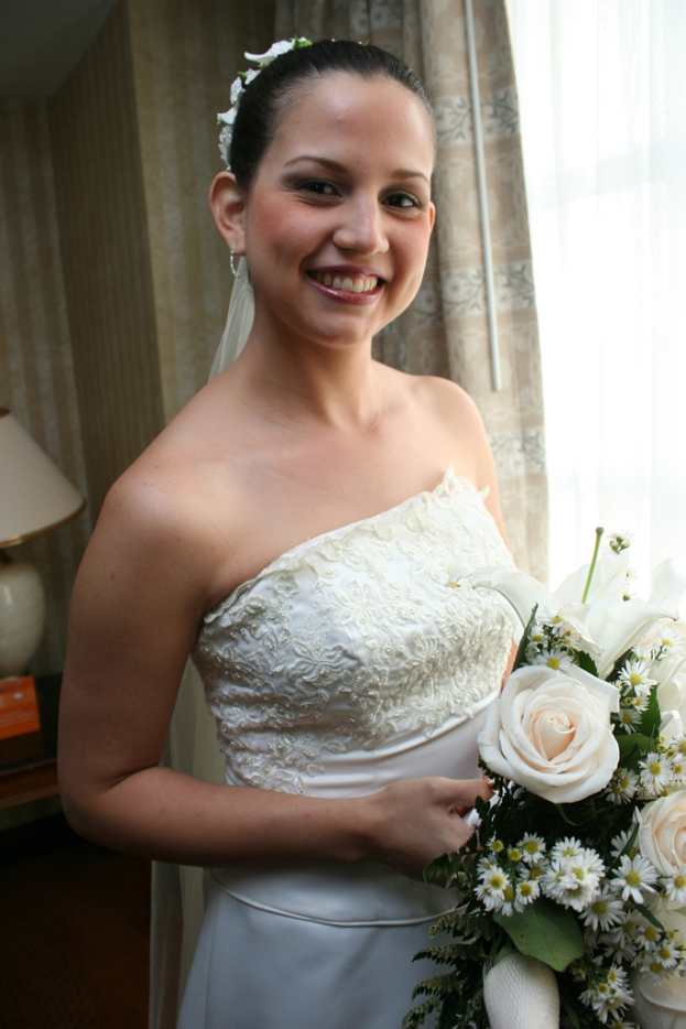 Eine strahlende Braut mit einem Blumenstrauß, vor einem Vorhang und einer Wand stehend und eine Lampe auf einem Tisch links im Bild.