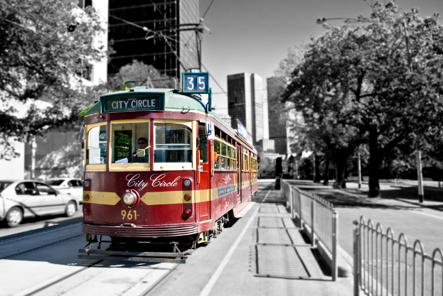 Ein roter Zug mit einer Person darin und Text darauf befindet sich in der Mitte, mit Autos, Bäumen, Gebäuden, Laternen und Drähten links und Bäumen und einem weißen Zaun rechts, mit Gebäuden und einer Person im Hintergrund.