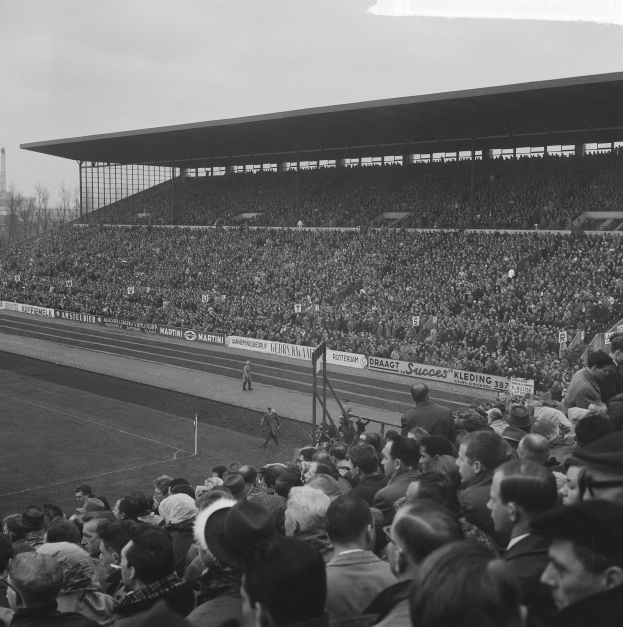 Schwarzes und weißes Foto einer vollen Tribüne mit Zuschauern bei einem Fußballspiel, mit Bannern, Pfählen, einem Schuppen, Bäumen, einem Turm und einem bewölkten Himmel.